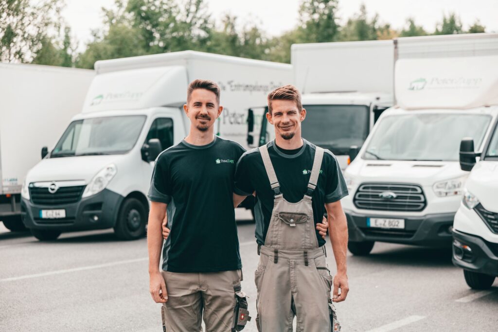 Zwei lächelnde Mitarbeiter des Entrümpelungsunternehmens Peter Zupp in Arbeitskleidung stehen Arm in Arm vor mehreren weißen Firmen-Transportern.